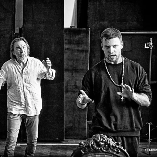 próba do spektaklu „Świętoszek", na zdjęciu: Bogusław Suszka z wyciągniętym palcem wskazującym, stojącym w tle oraz na pierwszym planie Łukasz Stawowczyk z różańcem na szyi, fot. Marcin Nowak. Zdjęcie jest czarno-białe