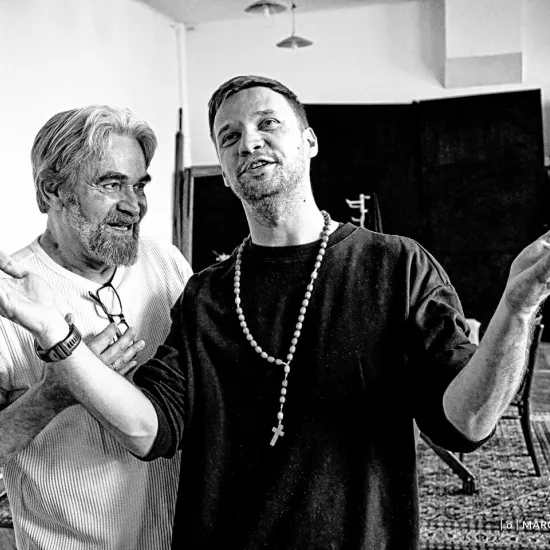 próba do spektaklu „Świętoszek", na zdjęciu: Dariusz Bereski stoi za Łukaszem Stawowczykiem. Łukasz ma rozłożone w modlitewnym geście ręce. Na szyi różaniec. Fot. Marcin Nowak, zdjęcie jest czarno-białe