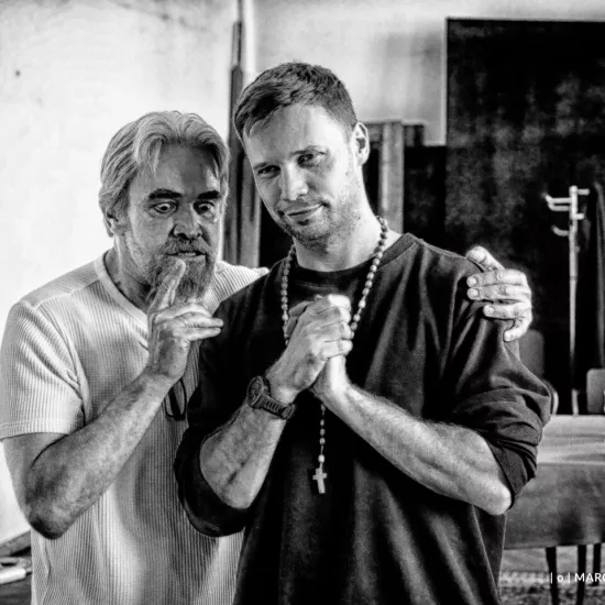 próba do spektaklu „Świętoszek", na zdjęciu: Dariusz Bereski, który stoi za drugim aktorem i szepcze mu coś do ucha i Łukasz Stawowczyk, który ma złożone dłonie jak do pacierza, fot. Marcin Nowak. Zdjęcie jest czarno-białe