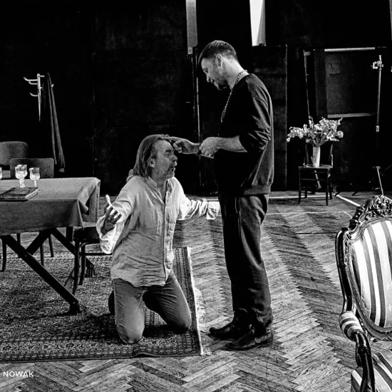 próba do spektaklu „Świętoszek", na zdjęciu: Bogusław Suszka, który klęczy i Łukasz Stawowczyk wykonujący gest błogosławieństwa na jego czole, fot. Marcin Nowak. Zdjęcie jest czarno-białe