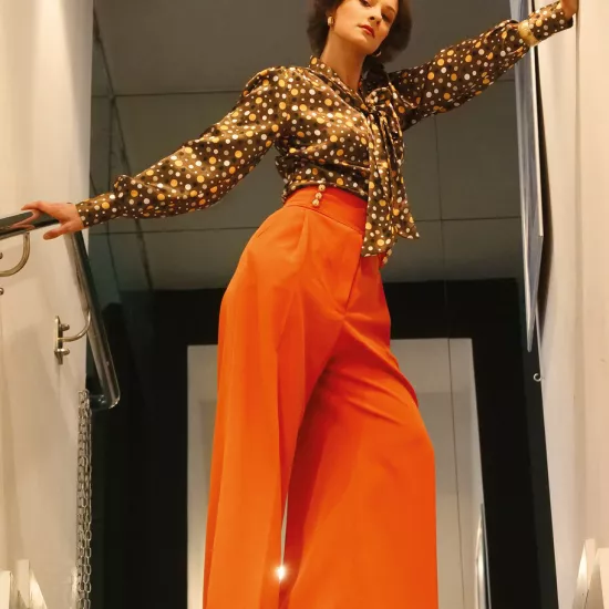 sesja zdjęciowa do spektaklu, na zdjęciu: Natalia Klepacka, która stoi na schodzach. Zdjęcie jest zrobione z dołu. Aktorka ma rozłożone ręce tak że dotyka ścian z obu stron, sylwetka jest skierowana w bok, twarz na wprost. Jest ubrana w pomarańczowe spodnie z rozszerzanymi nogawkami i wysokim stanem oraz elegancką bluzkę w brązowo-pomarańczowe kropki, fot. Marcin Nowak