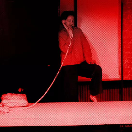 próba do spektaklu „Morderstwo w hotelu", zdjęcie zostało zrobione w sali prób, na zdjęciu: Natalia Klepacka, która stoi za łóżkiem i rozmawia przez telefon stacjonarny, którego słuchawka jest na sprężynowym, długim kablu, fot. Marcin Nowak. Na zdjęcie nałożony jest czerwony filtr. 