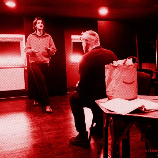 próba do spektaklu „Morderstwo w hotelu", zdjęcie zostało zrobione w sali prób, na zdjęciu: Natalia Klepacka idąca w kierunku siedzącego na krześle Mariusza Witkowskiego. Na niskim stoliku stoi papierowa torba z jedzeniem, obok leży scenariusz przedstawienia, fot. Marcin Nowak. Na zdjęcie nałożony jest czerwony filtr.