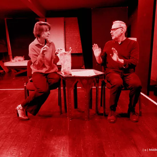 próba do spektaklu „Morderstwo w hotelu", zdjęcie zostało zrobione w sali prób, na zdjęciu: Natalia Klepacka, Mariusz Witkowski. Aktorzy siedzą na krzesłach przy niskim stoliku. Natalia Klepacka wyjmuje jedzenie z papierowej torby, fot. Marcin Nowak. Na zdjęcie nałożony jest czerwony filtr.