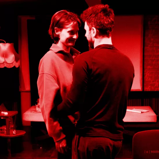 próba do spektaklu „Morderstwo w hotelu", zdjęcie zostało zrobione na sali prób,na zdjęciu: Natalia Klepacka, Krzysztof Wach. Aktorzy stoją blisko siebie, twarzami zwróconymi do siebie, obejmując się, fot. Marcin Nowak. Na zdjęcie nałożony jest czerowny filtr. 