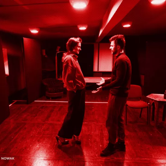 próba do spektaklu „Morderstwo w hotelu", zdjęcie zostało zrobione w sali prób,na zdjęciu: Natalia Klepacka, Krzysztof Wach. Aktorzy stoją od siebie w odległości, twarzami zwróconymi do siebie, trzymają się za ręce, fot. Marcin Nowak. Na zdjęcie nałożony jest czerowny filtr. 