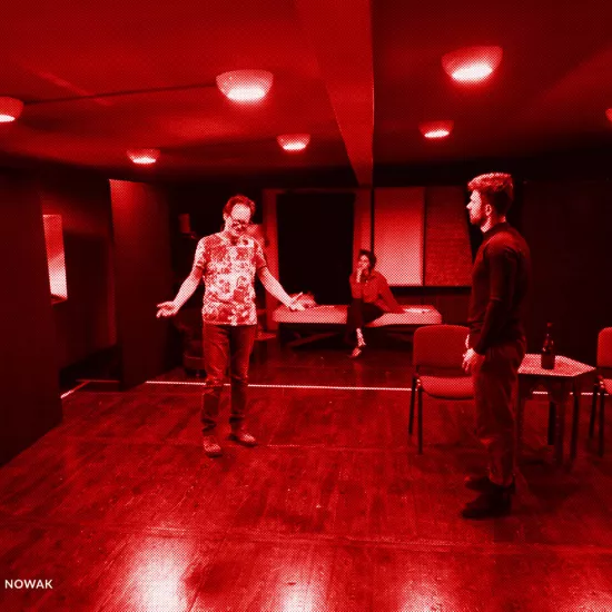 próba do spektaklu „Morderstwo w hotelu", zdjęcie z sali prób, na zdjęciu: Jacek Bończyk - reżyser spektaklu, który stoi w centralnym punkcie z rozłożonymi, skierowanymi w dół rękoma, z tyłu na łóżku siedzi w pozycji noga na nogę Natalia Klepacka, a przed reżyserem stoi Krzysztof Wach, po swojej prawej stronie ma dwa krzesła i niski stół, fot. Marcin Nowak. Na zdjęcie nałożony jest czerwony filtr.