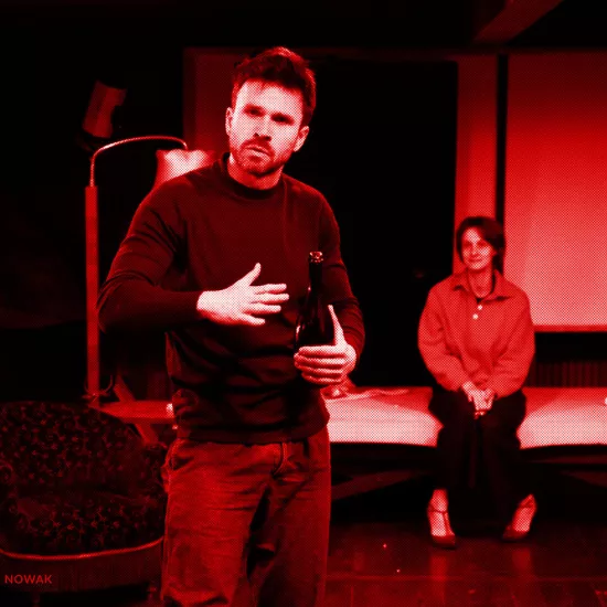 próba do spektaklu „Morderstwo w hotelu", zdjęcie zostało zrobione w sali prób, na zdjęciu: Krzysztof Wach trzymający w ręku butelkę szampana, w tle na łózku siedzi uśmiechnięta Natalia Klepacka, fot. Marcin Nowak. Na zdjęcie został nałożony czerwony filtr.