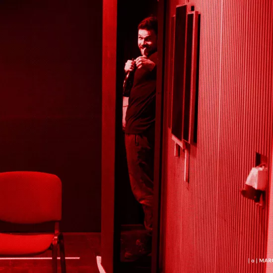 próba do spektaklu „Morderstwo w hotelu", zdjęcie zostało zrobione w sali prób, na zdjęciu: Krzysztof Wach wyglądający zza zastawki, aktor uśmiecha się, w prawej ręce trzyma butelkę szampana, a drugą pokazuje kciuk do góry, fot. Marcin Nowak. Na zdjęcie nałożony jest czerwony filtr.