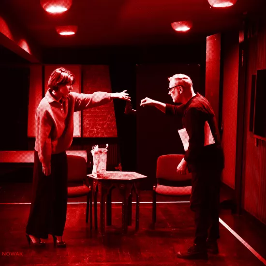 próba do spektaklu „Morderstwo w hotelu", zdjęcie zrobione w sali prób, na zdjęciu: Natalia Klepacka, która w palcach lewej dłoni trzyma pistolet i wręcza go z odległości Mariuszowi Witkowskiemu, fot. Marcin Nowak. Na zdjęcie został nałożony czerwony filtr.