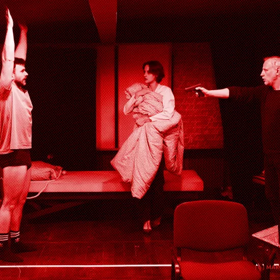 próba spektaklu „Morderstwo w hotelu", zdjęcie zostało wykonane w sali prób, na zdjęciu Krzysztof Wach, który ma na sobie koszulkę i bokserki, stoi z podniesionymi do góry rękoma, Natalia Klepacka stoi trzymając w ręku kołdrę oraz Mariusz Witkowski celujący pistoletem do Krzysztofa Wacha, fot. Macin Nowak. Na zdjęcie został nałożony czerwony filtr.