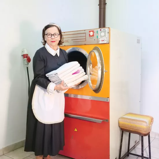 sesja zdjęciowa do spektaklu, na zdjęciu: Bogusława Pawelec w stroju pokojówki. Stoi przy dużej pralce, w dłoniach trzyma złożone pranie, spomiędzy ręczników wystaje buteleczka. Fot. Marcin Nowak