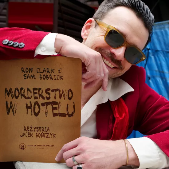 sesja zdjęciowa do spektaklu, na zdjęciu: Krzysztof Wach, który opiera się o książkę na której jest napis Morderstwo w hotelu. Aktor ma na sobie czerwoną marynarkę i białą koszulę. Na oczach przeciwsłoneczne, okrągłe okulary. Fot. Marcin Nowak