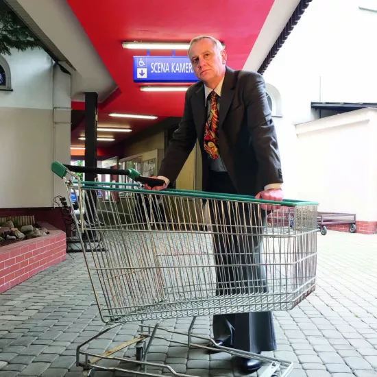 sesja zdjęciowa do spektaklu, na zdjęciu: Mariusz Witkowski stoi nad wózkiem sklepowym z pistoletem w ręku. Zdjęcie zrobione jest na podwórku, w przejściu na Scenę Kameralną. Na aktorem widnieje tablica podświetlana z napisem Scena Kameralna, fot. Marcin Nowak