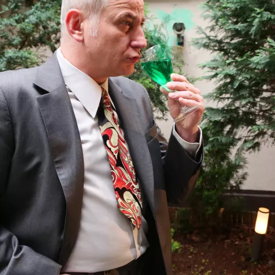 sesja zdjęciowa do spektaklu, na zdjęciu: Mariusz Witkowski ubrany w garnitur i kolorowy krawat, w dłoni trzyma kieliszek z zielonym płynem, drugą rękę trzyma w kieszeni, fot. Marcin Nowak