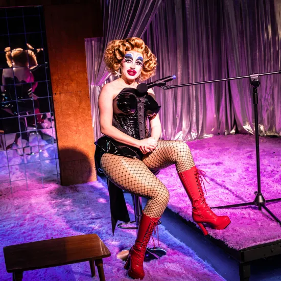 Zdjęcie ze spektaklu „Czas na wielki numer". Na fotografii widać aktorkę Matyldę Paszczenko ucharakteryzowaną na drag queen. Aktorka siedzi na stołku typu hoper. Do tworzy ma skierowany mikrofon. Ma na sobie czerwone, długie, skórzane kozaki na wysokim obsacie i lateksowy strój a la body. Aktorka ma wypchane uda i biodra. Ma na sobie także dużą, brązową perukę. Jej makijaż jest ostry, wyrazisty, wychodzący poza kontury twarzy. Autorką fotografii jest Natalia Kabanow 