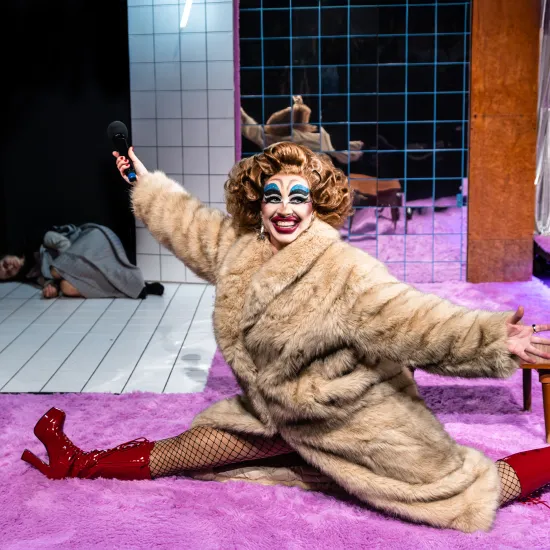 Zdjęcie ze spektaklu „Czas na wielki numer". Na fotorafii widać aktorkę Matyldę Paszczenko w charakteryzacji na drag queen. Ma na sobie gruby płaszcz. Robi szpagat. W tle pod drzwiami leży aktorka Natalia Klepacka. Autorką zdjęcią jest Natalia Kabanow