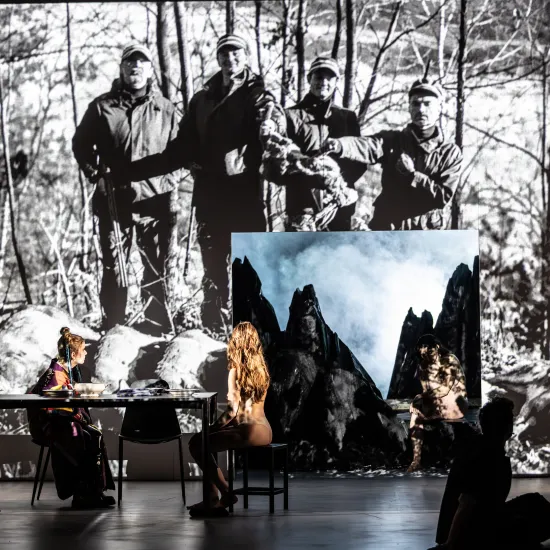Zdjęcie ze spektaklu „Prowadź swój pług przez kości umarłych". Na Dużej Scenie Teatr Jaracza widać długi stół przy którym siedzą dwie tancerki i aktorka Sara Lityńska, patrzą się na wyświetlane na ścianie zdjęcie myśliwych, w ścianie jest kwadratowa dziura przez którą widać góry. Obok dziury stoi aktorka Milena Lisiecka. Autorką zdjęcia jest Natalia Kabanow