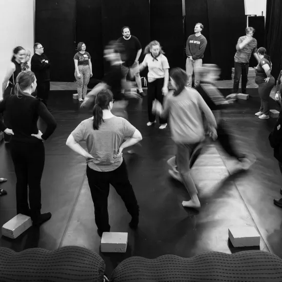 Czarno-białe zdjęcie przedstawiające grupę osób w sali prób. Uczestnicy są ustawieni w szerokim kręgu na ciemnej podłodze, a przed wieloma z nich leżą jasne, prostokątne klocki. Część osób stoi nieruchomo, obserwując środek kręgu, gdzie dwie postacie są rozmyte w ruchu, co sugeruje dynamikę ćwiczenia lub zabawy ruchowej. W tle widać proste ściany, rzędy krzeseł oraz ciemne kotary. Atmosfera wydaje się skupiona i warsztatowa.