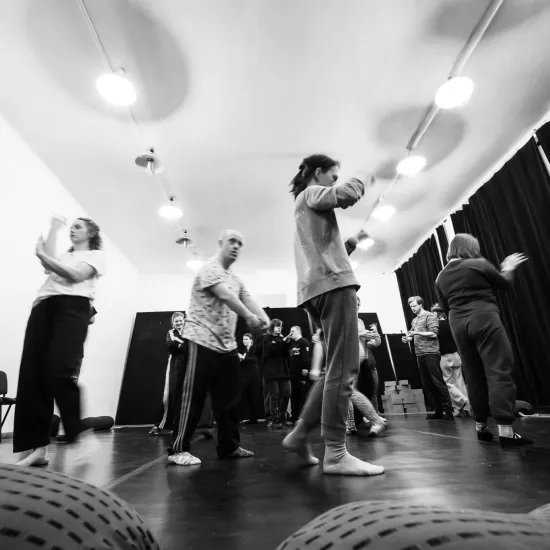 Czarno-białe zdjęcie wykonane z niskiej perspektywy (z poziomu podłogi), przedstawiające grupę osób podczas dynamicznych warsztatów ruchowych w sali z ciemną podłogą i białym sufitem. Wiele postaci jest rozmytych, co podkreśla ekspresję i tempo ćwiczeń. Uczestnicy wykonują różnorodne gesty rękami i są w trakcie przemieszczania się po przestrzeni. Na suficie widać rzędy jasnych, okrągłych lamp, a w tle czarną kotarę i rządy krzeseł. Na pierwszym planie widoczne są fragmenty wypukłych, fakturowanych puf.