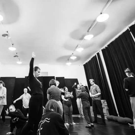 Czarno-białe zdjęcie dokumentujące warsztaty artystyczne w sali z ciemną podłogą i jasnym sufitem. Grupa kilkunastu osób jest rozproszona po przestrzeni, zastygnięta w różnych pozach. Na pierwszym planie po lewej stronie stoi kobieta z wysoko uniesioną jedną ręką, a obok niej inna osoba kuca w dynamicznym gestem. Pozostali uczestnicy są widoczni w głębi sali – niektórzy stoją, inni wykonują gesty dłońmi. W tle widać rzędy jasnych lamp na suficie oraz czarne kotary przesłaniające ściany.