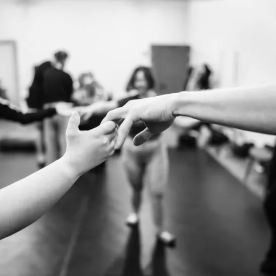 Czarno-białe zdjęcie o płytkiej głębi ostrości, przedstawiające zbliżenie na dłonie dwóch osób, które delikatnie stykają się palcami w geście porozumienia lub wspólnego ruchu. Punkt ostrości jest ustawiony na dłoniach znajdujących się w centrum kadru. W rozmazanym, jasnym tle widać sylwetki pozostałych uczestników warsztatów, w tym uśmiechniętą kobietę po lewej stronie. Scena oddaje atmosferę bliskości, zaufania i emocjonalnego zaangażowania podczas zajęć grupowych w sali prób.