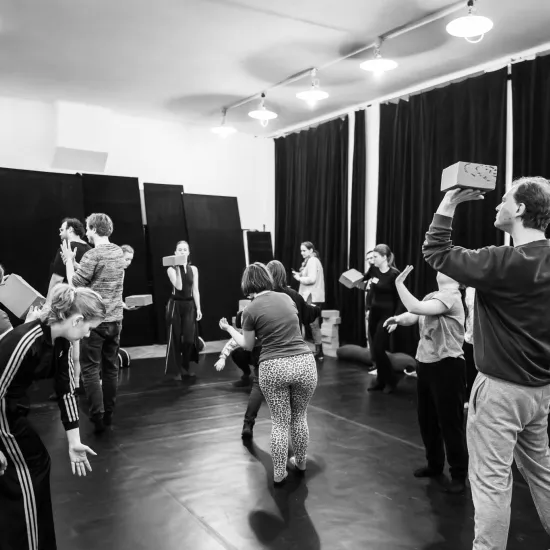 Czarno-białe zdjęcie przedstawiające grupę osób podczas ćwiczeń warsztatowych w sali z ciemną podłogą i czarnymi kotarami. Uczestnicy wchodzą w interakcje z jasnymi, prostokątnymi klockami – niektórzy trzymają je w dłoniach, inni balansują nimi na głowie lub ramieniu, a jeszcze inni wykonują gesty bez klocków. Na pierwszym planie po prawej stronie mężczyzna stoi profilem, trzymając klocek na dłoni przy głowie. Po lewej stronie kobieta w dresie z paskami jest pochylona w dynamicznej pozie.