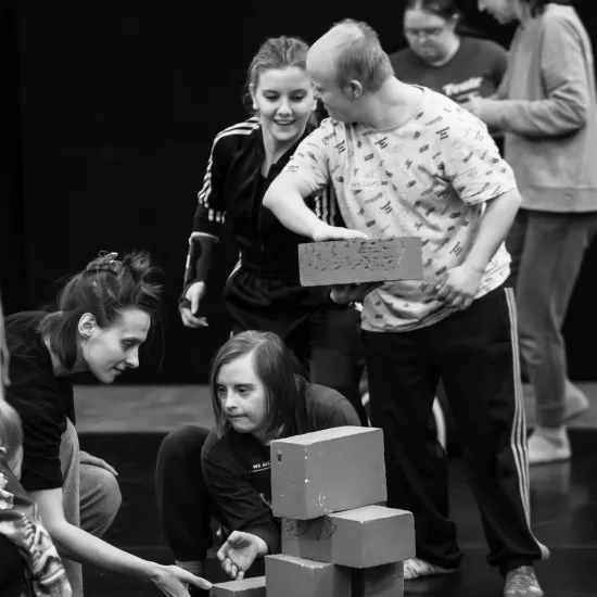 Czarno-białe zdjęcie w pionowym kadrze, przedstawiające grupę osób wspólnie budujących konstrukcję z jasnych, prostokątnych klocków na ciemnej podłodze. W centrum uwagi znajduje się mężczyzna w jasnej koszulce, który precyzyjnie dokłada klocek na szczyt wieży. Obok niego uśmiechnięta kobieta w sportowej bluzie z uwagą śledzi jego ruchy. Dwie inne osoby kucają przy podstawie budowli, a jedna z nich wyciąga dłoń, by podtrzymać lub skorygować ustawienie dolnego elementu.