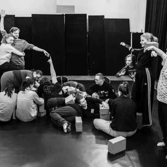 Czarno-białe zdjęcie panoramiczne przedstawiające grupę osób tworzącą wspólną, wielopoziomową rzeźbę z własnych ciał i jasnych klocków na środku sali. Część osób leży lub siedzi na podłodze, podczas gdy inne stoją, tworząc wyższe punkty kompozycji. Po lewej stronie mężczyzna i kobieta splatają ramiona, unosząc dłonie w górę. Po prawej stronie uczestnicy balansują klockami na barkach i wyciągają ręce w bok. Całość tworzy dynamiczną, geometryczną strukturę na tle czarnych paneli i kotar.