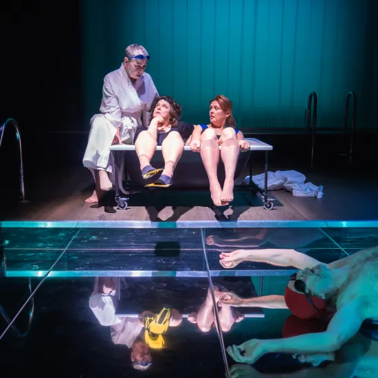 Scena ze spektaklu „Pływalnia” w Teatrze Jaracza, podzielona na dwa plany. Na podwyższeniu w głębi sceny trzy aktorki – jedna w białym szlafroku, dwie w strojach kąpielowych – siedzą ściśnięte w czarnej wannie na kółkach i rozmawiają. Na pierwszym planie, na błyszczącej, lustrzanej podłodze imitującej taflę wody, leży na plecach aktor w czerwonych kąpielówkach i czepku. Całość oświetlona jest chłodnym, turkusowym światłem, a z boku widoczne są drabinki basenowe.