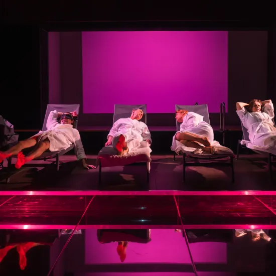 Scena ze spektaklu „Pływalnia” w Teatrze Jaracza. Czworo aktorów w białych szlafrokach leży obok siebie na leżakach basenowych ustawionych w głębi sceny. Całość tonie w intensywnym, różowo-fioletowym świetle reflektorów. Na pierwszym planie znajduje się gładka, ciemna podłoga, w której odbijają się postacie i zarysy leżaków, a po lewej stronie widać metalową drabinkę basenową.