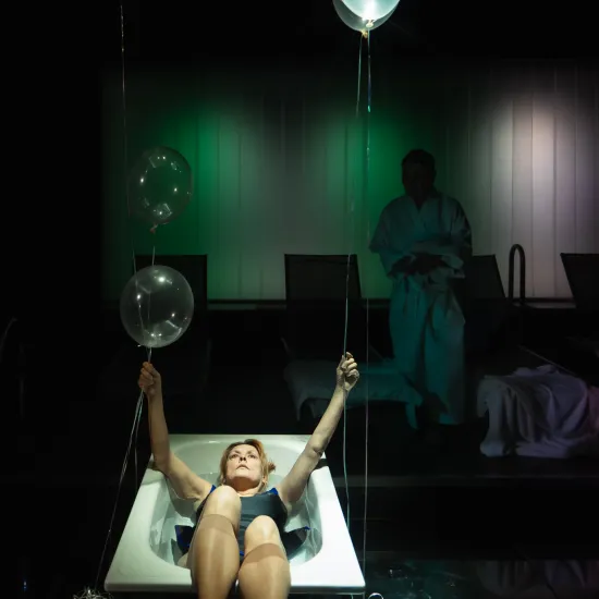Kadr ze spektaklu „Pływalnia” w Teatrze Jaracza. W centrum uwagi znajduje się aktorka w ciemnym stroju kąpielowym, która leży w białej wannie ustawionej na scenie. Kobieta patrzy w górę, trzymając w uniesionych dłoniach sznurki od kilku przezroczystych, świecących balonów, które unoszą się nad nią. W ciemnym tle, oświetlonym z tyłu zielonym światłem, stoi inna postać w białym szlafroku oraz widoczne są zarysy leżaków basenowych.