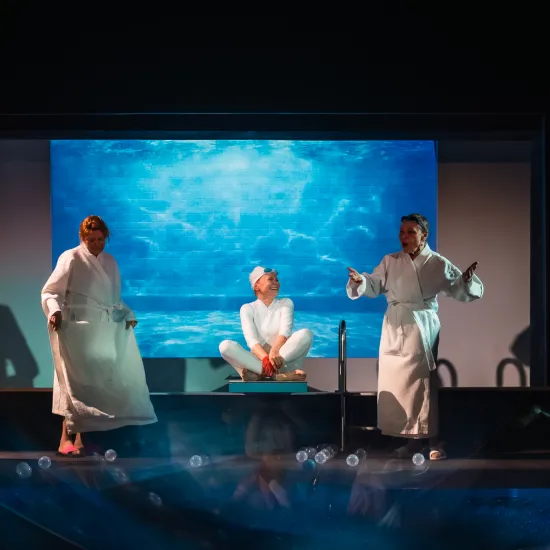 Scena ze spektaklu „Pływalnia” w Teatrze Jaracza. W centrum podwyższenia siedzi aktorka w białym stroju i czepku, otoczona przez czworo innych aktorów w białych szlafrokach, którzy żywo gestykulują. W tle widać duży ekran z podwodną, błękitną grafiką basenu, a na pierwszym planie rozsypane, małe kulki.