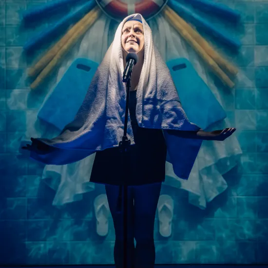 Kadr ze spektaklu „Pływalnia” w Teatrze Jaracza. W centrum stoi aktorka w ciemnym stroju kąpielowym z dużym, białym ręcznikiem narzuconym na głowę niczym welon lub chusta. Kobieta ma uniesioną głowę, patrzy w górę z natchnioną miną i stoi przed mikrofonem na statywie, rozkładając ręce w boki. Bezpośrednio nad jej głową, na tle ściany z niebieskich kafelków basenowych, wisi koło ratunkowe oraz żółte i niebieskie makarony do pływania, tworzące wokół niej wzór przypominający aureolę.