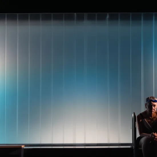 Scena ze spektaklu „Pływalnia” w Teatrze Jaracza. W prawym dolnym rogu kadru siedzi samotna aktorka w ciemnym stroju kąpielowym i goglach na głowie, mając splątane dłonie i opuszczony wzrok. Tło zdjęcia stanowi duża, minimalistyczna, pionowo prążkowana ściana oświetlona chłodnym, błękitno-białym światłem reflektorów, co tworzy atmosferę pustki i melancholii. Obok kobiety widoczna jest metalowa drabinka basenowa.