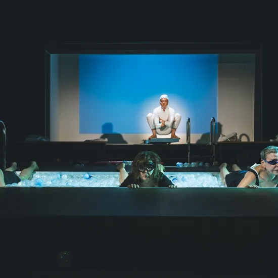Scena ze spektaklu „Pływalnia” w Teatrze Jaracza. Na pierwszym planie trzy aktorki w strojach kąpielowych i goglach leżą w podłużnym basenie wypełnionym białymi i błękitnymi kulkami, opierając się o krawędź sceny. W głębi, na podwyższeniu, siedzi w kucki czwarta aktorka w białym stroju i czepku, oświetlona na tle jasnoniebieskiego ekranu. Po prawej stronie w ciemności widać siedzącego aktora, a po lewej metalową drabinkę basenową.