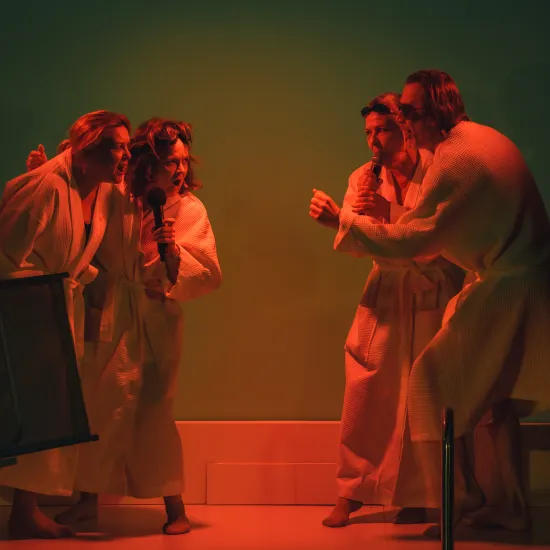 Scena ze spektaklu „Pływalnia” w Teatrze Jaracza. Czworo aktorów ubranych w białe szlafroki kąpielowe stoi na scenie, podzielonych na dwie ekspresyjne pary stojące naprzeciw siebie. Dwie osoby trzymają mikrofony, a wszyscy mają dynamiczne, pełne emocji miny, jakby śpiewali lub krzyczeli. Całość oświetlona jest intensywnym, kontrastowym światłem – czerwonym od dołu i zielonym w tle, co nadaje scenie dramatyczny charakter. Po lewej stronie widoczny jest przewrócony leżak, a po prawej fragment drabinki basenow